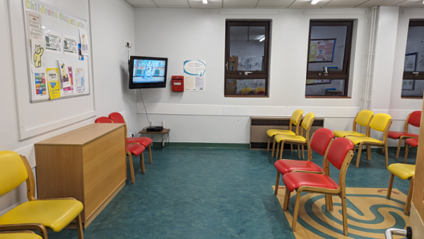 Image shows the corner of the children’s waiting room with pairs of red and yellow chairs placed around the room. There is an information board covered in posters next to a TV playing cartoons.