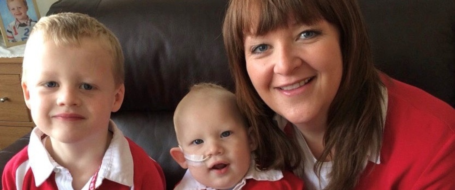 A family photo of Zachary Bowen as a baby, wearing his NG tube, with brother Harrison and mum Sarah smiling at the camera.