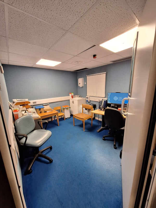 Image shows small office with the Audiologists desk in the corner and a wooden patient’s chair beside it.