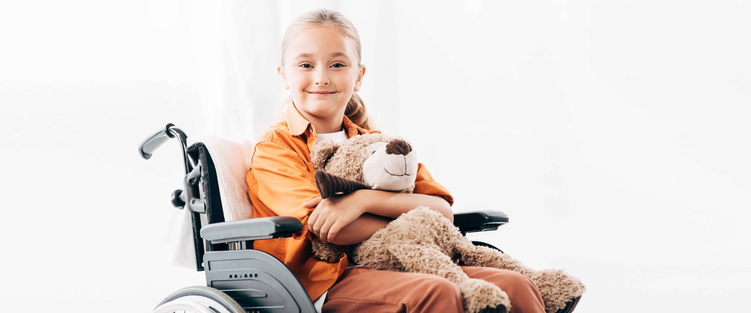 Image shows girl in wheelchair smiling and holding a teddy bear.