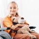 Image shows girl in wheelchair smiling and holding a teddy bear.