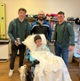 Image shows three men standing around a woman in a hospital bed