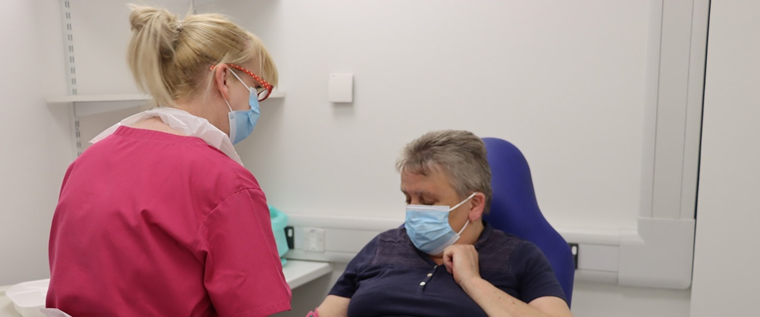 Image shows Mary sat in a chair while blood is drawn by staff member Carol.