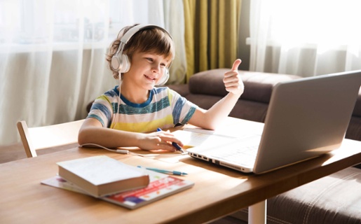 Image shows young boy sat in front of laptop smiling and holding up his thumb.