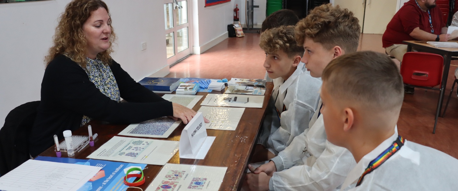 A scientist points to a worksheet with children in lab coats listening to what she is saying