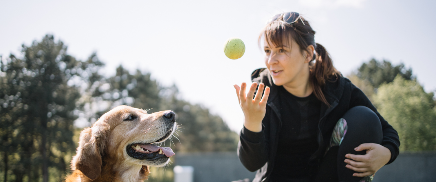 A woman is pictured sitting next to a dog and throwing a tennis ball.