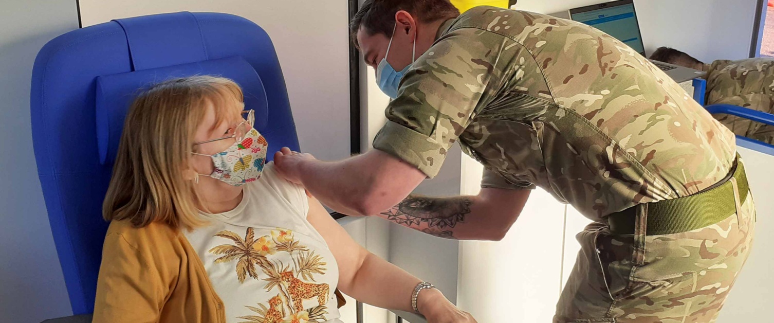 A woman sat in a chair is vaccinated in the arm by an Army medic.