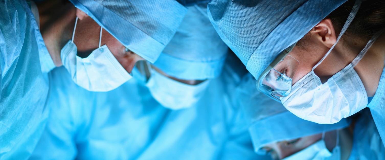 Three surgeons in blue scrubs and face masks lean over a patient.