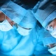 Three surgeons in blue scrubs and face masks lean over a patient.