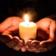 A pair of hands are cupped together, holding a small lit candle as darkness shrouds around the scene.