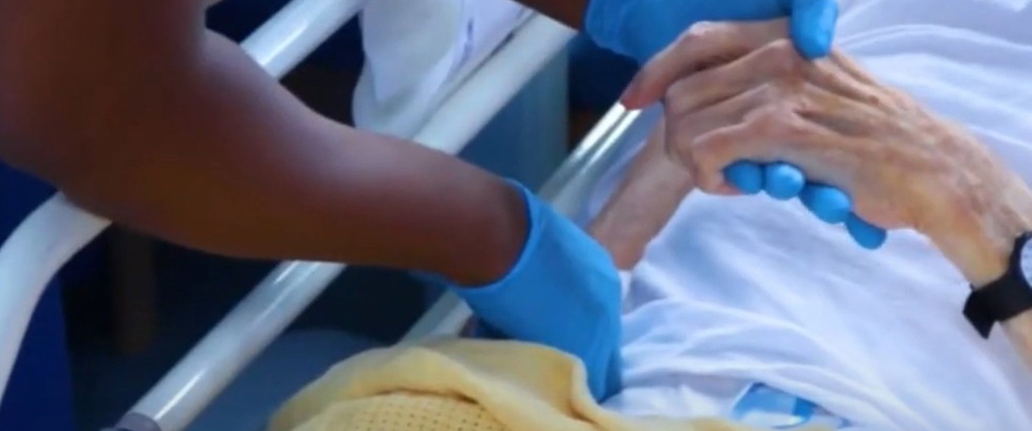 Image is a close-up of two people holding hands in a hospital setting.