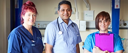 A nurse, a doctor and another nurse stand on a hospital ward smiling at the camera.