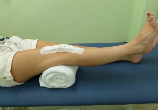 A patient is on a bed. Their right leg is raised with a rolled towel under their knee.