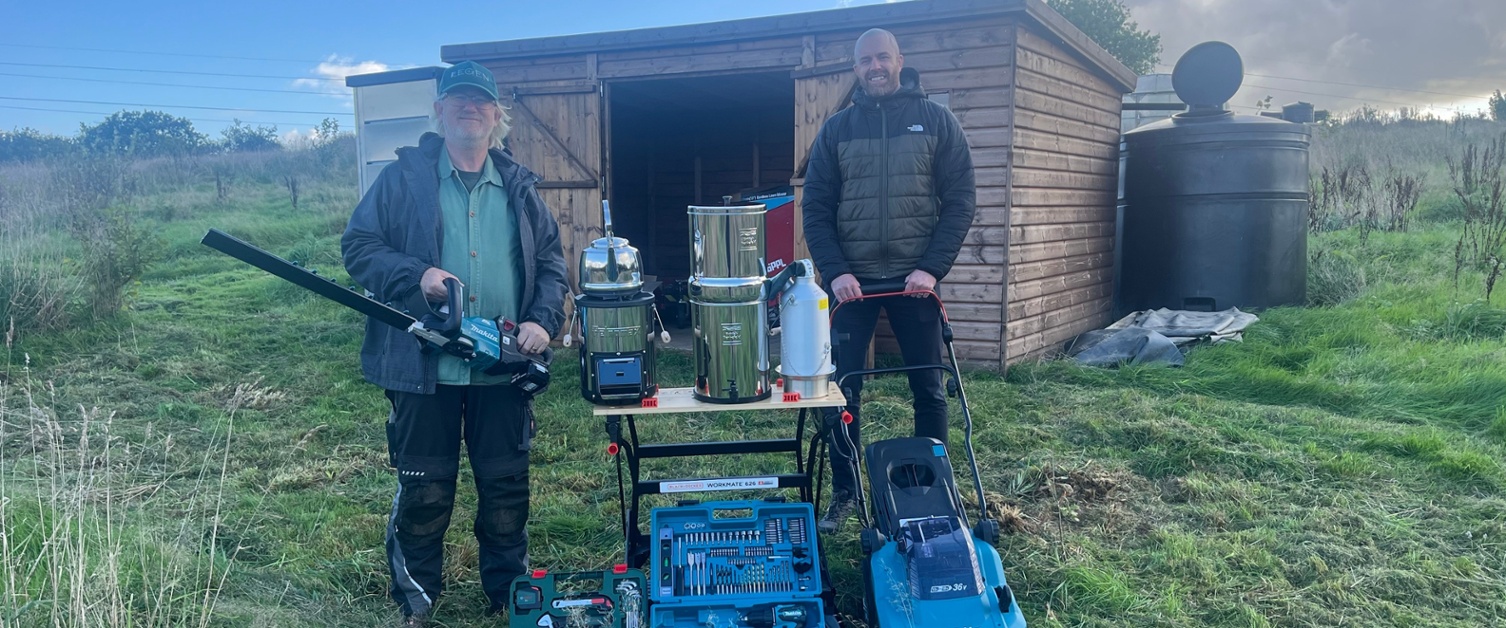 Image shows two men stood in front of a shed holding tools