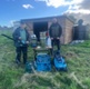 Image shows two men stood in front of a shed holding tools