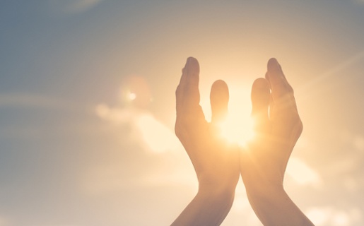 An image of hands being held up, with the sun in the distance shining through them.