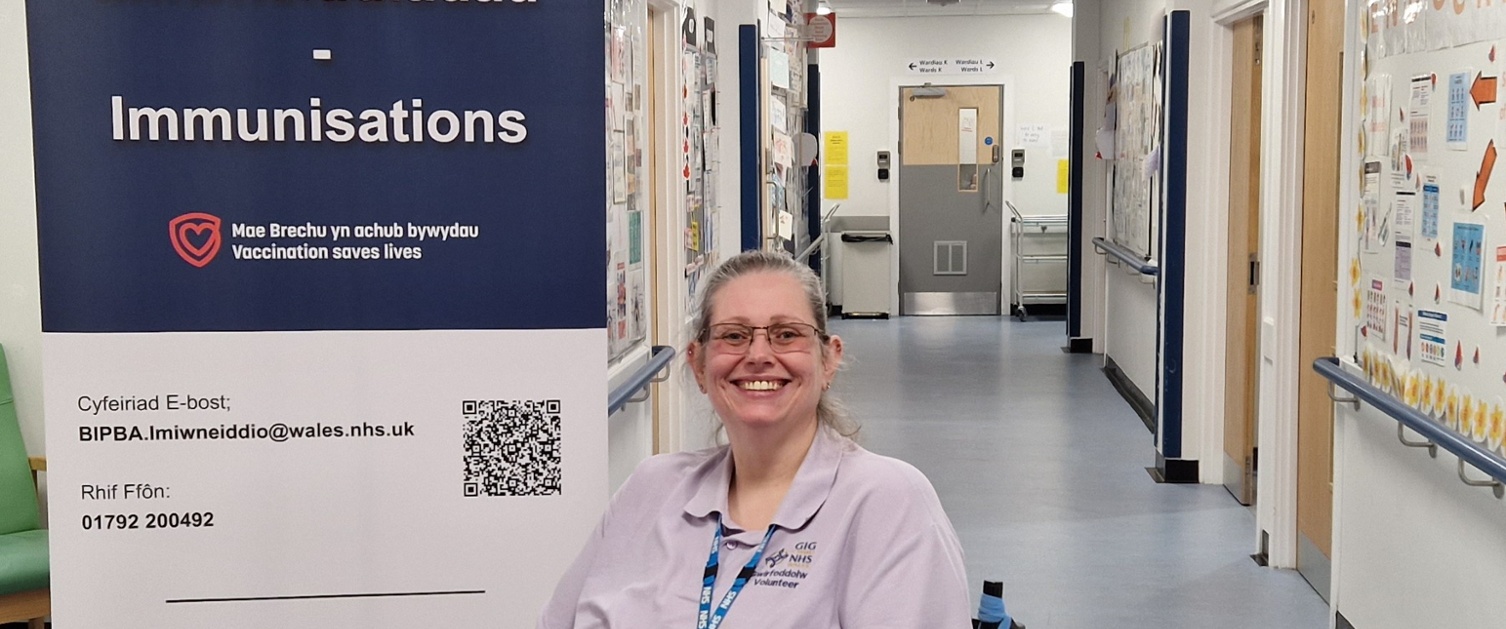 An image of a woman smiling at the camera, she is sat in a wheelchair in a hospital corridor.
