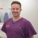 Image of Tom Henwood smiling at camera and wearing purple scrubs.