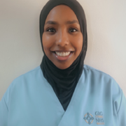 A young Muslim woman, wearing a black hijab and pale blue NHS scrubs, smiles at the camera.