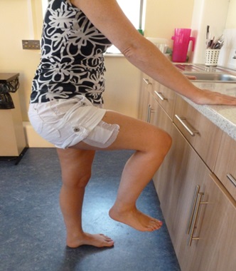 A patient stands facing a kitchen worktop. Their right leg is raised.