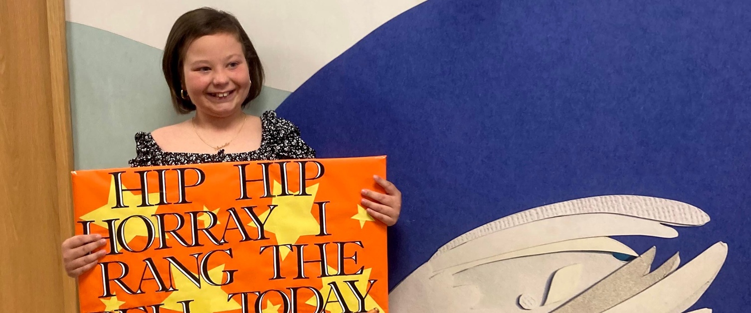 Isabella holds a sign saying Hip Hip Hooray I Rang The Bell Today.