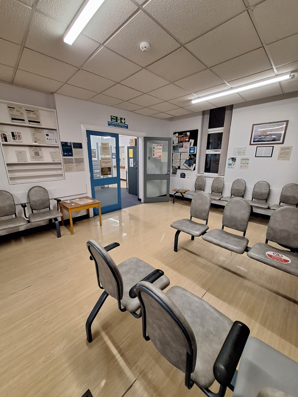 Image shows 4 rows of grey chairs in a large room with posters on the wall. There are double glass doors in the corner of the room leading to the outpatient department.