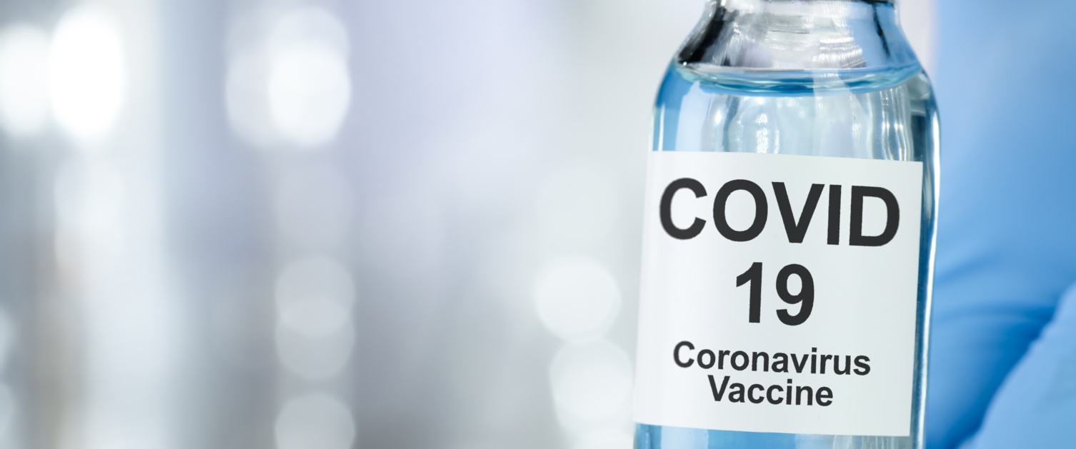 Close up of a vial labelled Covid vaccine held between two fingers on a gloved hand.