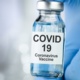 Close up of a vial labelled Covid vaccine held between two fingers on a gloved hand.