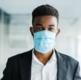 Image shows a young man in a suit facing the camera with a blue surgical mask on.