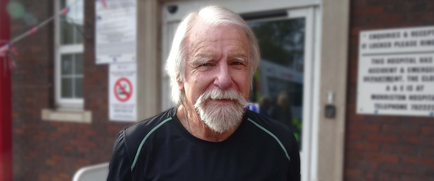 Image is of a man with white hair and beard standing outside a hospital.