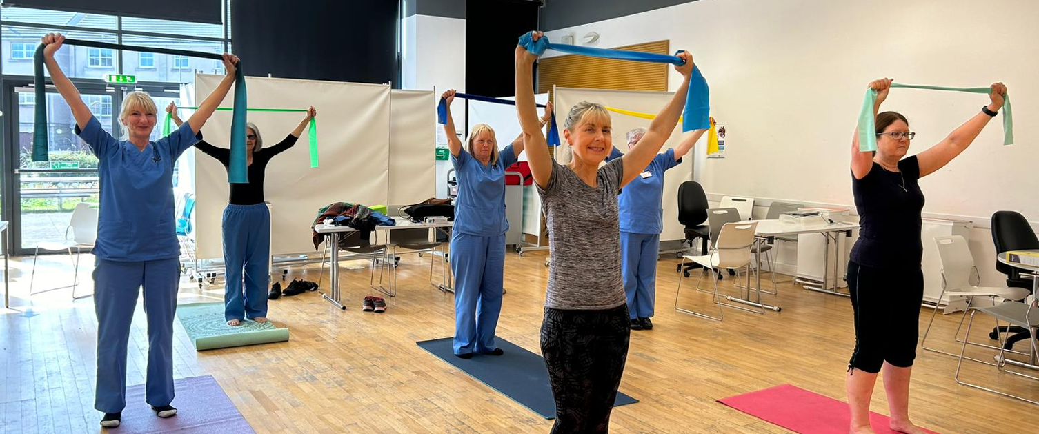 Staff members stand on mats with arms above their heads pulling a resistance band.