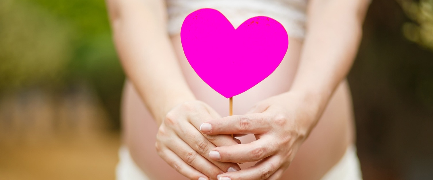 A pregnant woman holds a pink paper heart in front of her tummy.