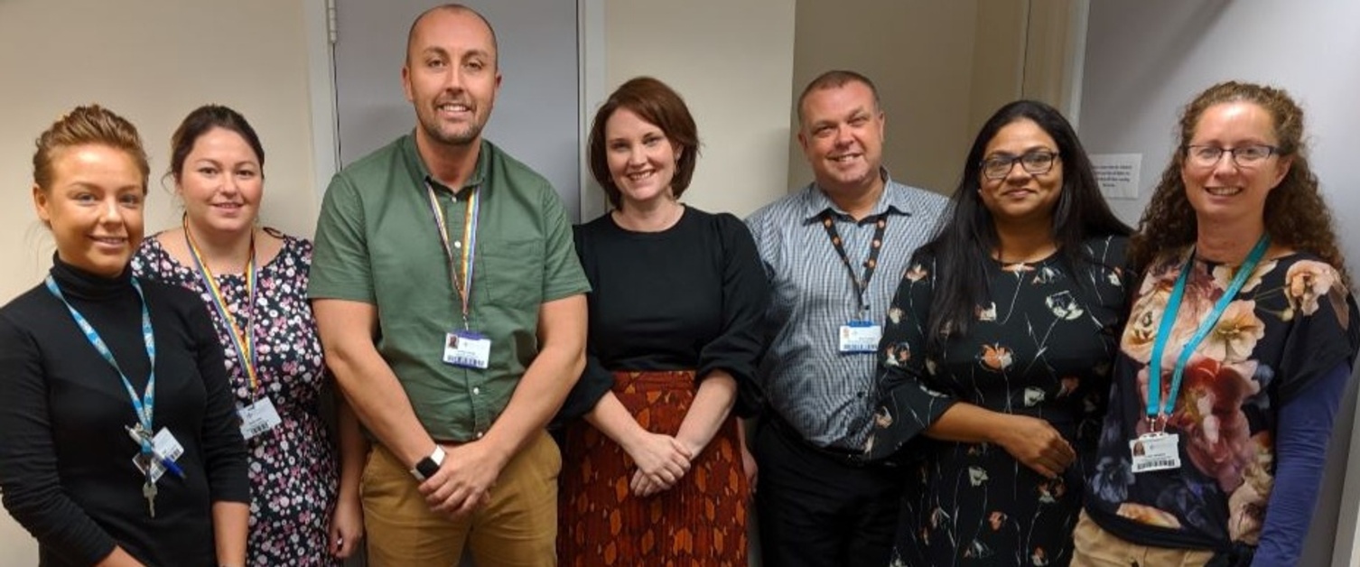 Seven members of staff from the Department of Liaison Psychiatry, stand in a office, smiling at the camera.