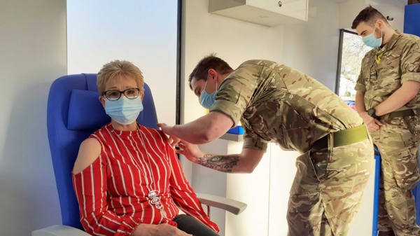 A woman, seated, is vaccinated in the arm by an Army medic.