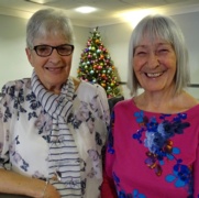 Volunteers Ann Humphrey and Linda Fisher smile in front of a Christmas tree.