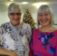 Volunteers Ann Humphrey and Linda Fisher smile in front of a Christmas tree.