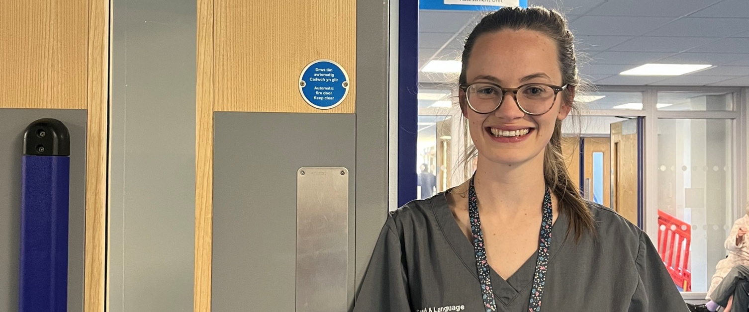 Image shows a woman wearing glasses and standing in a hospital corridor