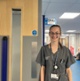 Image shows a woman wearing glasses and standing in a hospital corridor