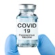 Gloved fingers holding a Covid vaccine vial plus a syringe in vial bottle.