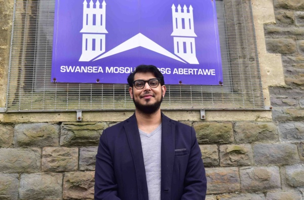 Farid Ali, wearing a navy jacket, stands outside in front of Swansea Mosque sign facing the camera.