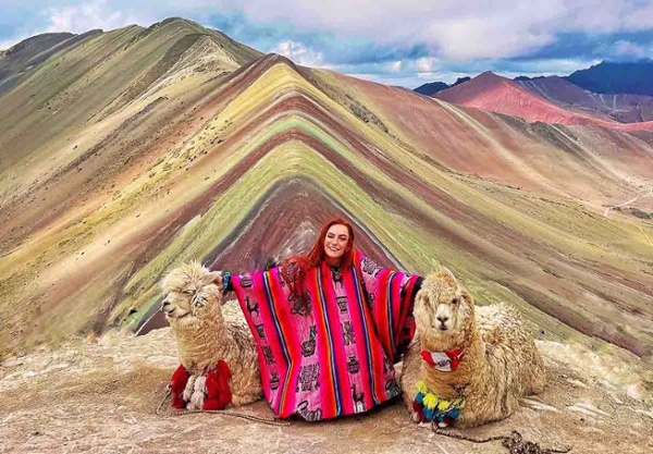 Image shows a woman with two alpacas in mountainous terrain.