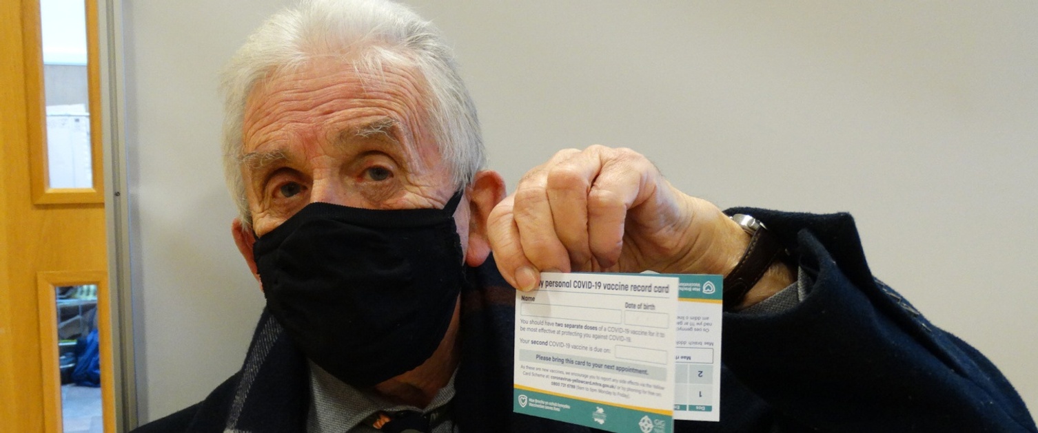 Image shows a man wearing a face mask holding up his vaccination card.