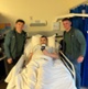 Image shows two men standing around a woman in a hospital bed