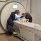Two scientists stand either side of an MRI scanner working on something inside.