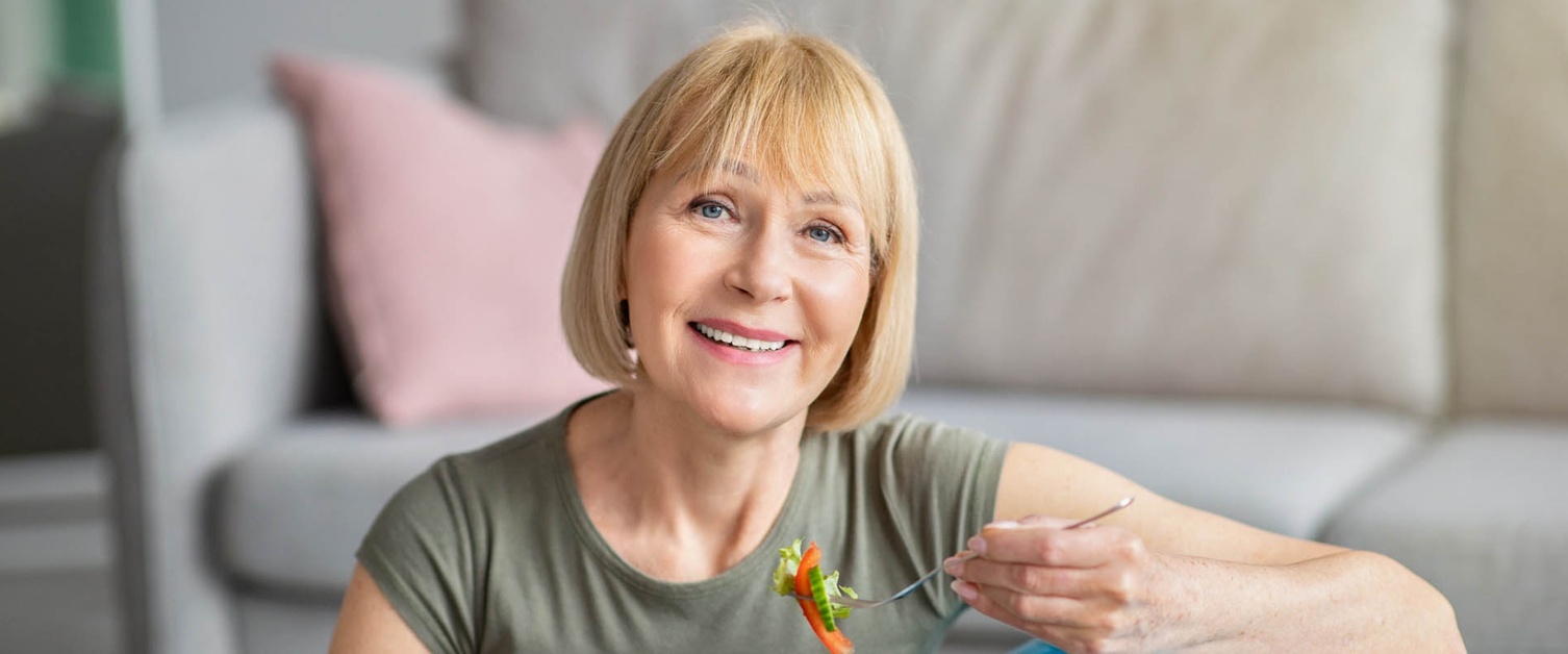 A woman sat on the floor leans against a gym ball eating a salad.