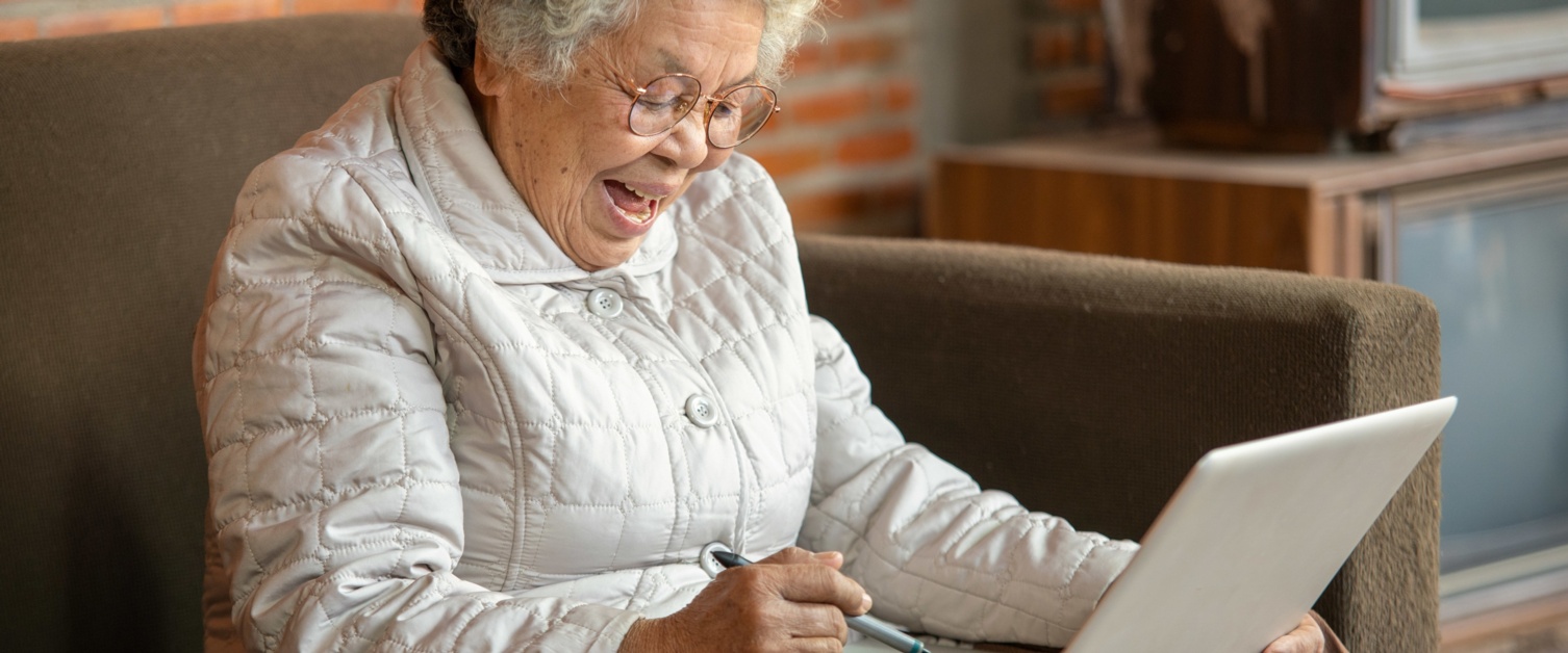 A pensioner sits on a sofa with a laptop on her lap. She is smiling.