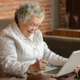 A pensioner sits on a sofa with a laptop on her lap. She is smiling.