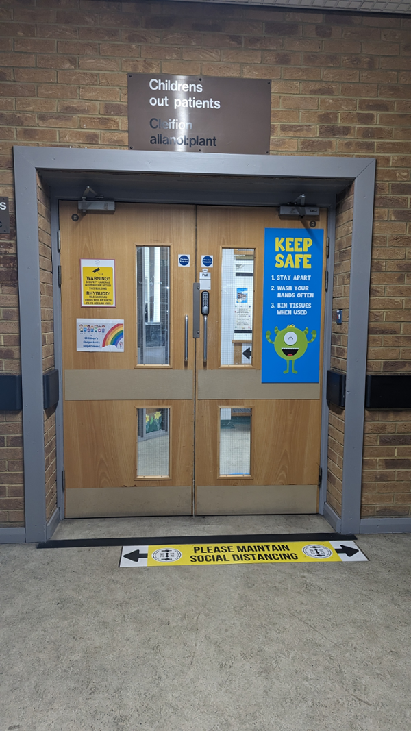 Image shows double wooden doors with a keypad lock in the middle. There is a sign above the door stating “Childrens Outpatients”.