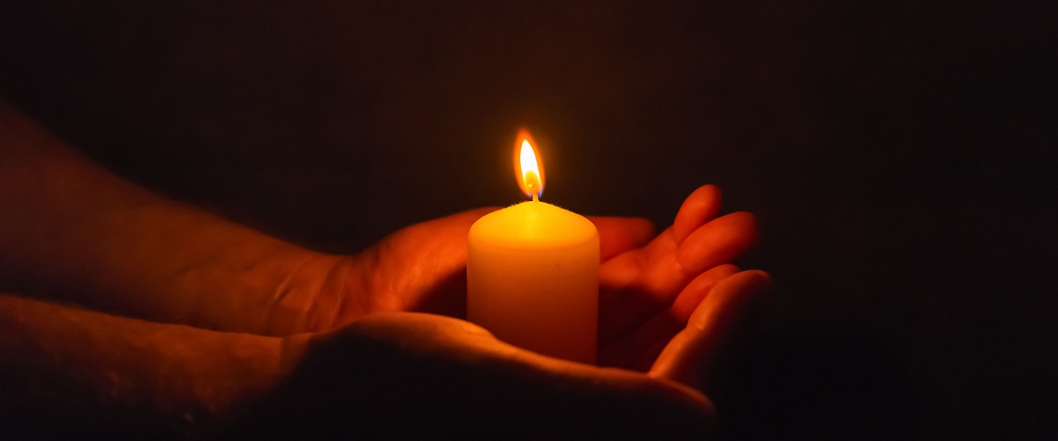 Image shows a lit candle upright being held in the palms of hands in a dark room.