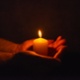 Image shows a lit candle upright being held in the palms of hands in a dark room.